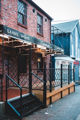 A brick building with decorative lattice windows and a wooden deck features a sign with multiple brand names. The building is adorned with string lights and has a metal railing leading up to a black stairway. Adjacent structures are visible, displaying a mix of brick and painted surfaces.
