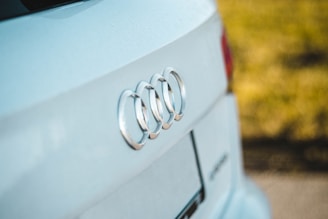 A close-up view of the rear of a white car, featuring the prominent chrome logo of a well-known automobile brand. The background is softly blurred, accentuating the sharpness of the car's emblem and its clean surface.