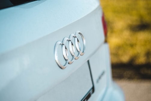 A close-up view of the rear of a white car, featuring the prominent chrome logo of a well-known automobile brand. The background is softly blurred, accentuating the sharpness of the car's emblem and its clean surface.