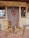 Two wooden rocking chairs are placed on a brick-patterned porch in front of a rustic door adorned with a festive wreath. The wreath is decorated with red berries, pine cones, and a large red bow. The building's exterior has a textured stucco finish with wooden-framed windows.