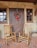 Two wooden rocking chairs are placed on a brick-patterned porch in front of a rustic door adorned with a festive wreath. The wreath is decorated with red berries, pine cones, and a large red bow. The building's exterior has a textured stucco finish with wooden-framed windows.