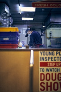A person wearing a cap and face mask is inside a kitchen or food preparation area, focused on their work. The setting includes shelves with colorful storage containers and various kitchen equipment. A sign with bold text, partially visible, suggests a promotional or instructional message.