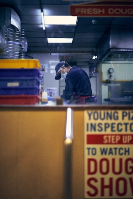 A person wearing a cap and face mask is inside a kitchen or food preparation area, focused on their work. The setting includes shelves with colorful storage containers and various kitchen equipment. A sign with bold text, partially visible, suggests a promotional or instructional message.
