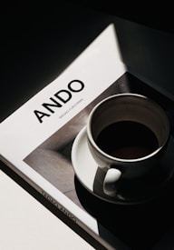 A hardcover book with the title 'ANDO' and the author's name, Masao Furuyama, is resting under the soft light. A filled coffee cup is placed on the book, casting a shadow that contrasts with the bright illumination.