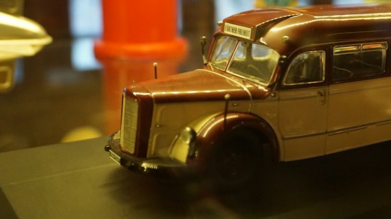 A detailed model of a vintage bus with an exterior painted in classic maroon and beige. The bus displays a sign reading 'Fürther Freiheit' and is placed on a dark platform. The background is slightly out of focus, featuring blurred objects including a red cylindrical shape.