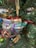 A festive ornament shaped like Santa Claus transformed into a mermaid, hanging on a Christmas tree. The Santa mermaid is adorned with vibrant colors, including reds, blues, and greens, and holds a small golden fish. The ornament is surrounded by green pine needles and colorful holiday lights.