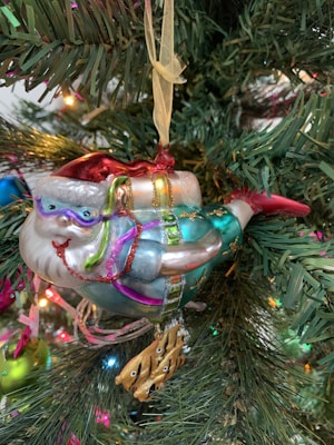 A festive ornament shaped like Santa Claus transformed into a mermaid, hanging on a Christmas tree. The Santa mermaid is adorned with vibrant colors, including reds, blues, and greens, and holds a small golden fish. The ornament is surrounded by green pine needles and colorful holiday lights.