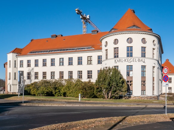 A large, historic building with a distinctive red-tiled roof and white facade, featuring multiple windows and a rounded corner with 'Karl-Kegel-Bau' written on it. The structure is situated on a corner with a crane visible in the background, and surrounded by greenery and a well-maintained lawn. Traffic signs indicating a speed limit and no parking are present in the foreground.