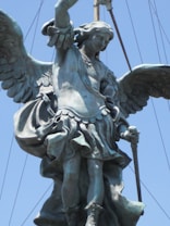 A large, detailed statue of an angel with expansive wings, holding a sword, crafted from metal. The figure is depicted in flowing, classical garments with attention to texture and motion, against a clear blue sky.