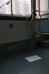 An empty section of public transport with a visible wheelchair accessibility sign on the wall. The interior has gray flooring, a railing painted in yellow, and a window showing an urban backdrop with tall apartment buildings. A piece of paper lies on the floor.