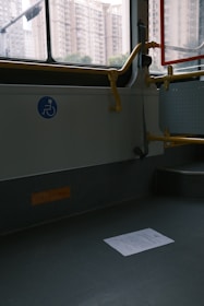An empty section of public transport with a visible wheelchair accessibility sign on the wall. The interior has gray flooring, a railing painted in yellow, and a window showing an urban backdrop with tall apartment buildings. A piece of paper lies on the floor.