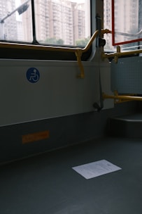 An empty section of public transport with a visible wheelchair accessibility sign on the wall. The interior has gray flooring, a railing painted in yellow, and a window showing an urban backdrop with tall apartment buildings. A piece of paper lies on the floor.