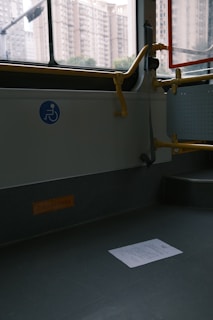 An empty section of public transport with a visible wheelchair accessibility sign on the wall. The interior has gray flooring, a railing painted in yellow, and a window showing an urban backdrop with tall apartment buildings. A piece of paper lies on the floor.