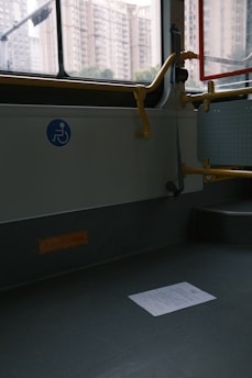 An empty section of public transport with a visible wheelchair accessibility sign on the wall. The interior has gray flooring, a railing painted in yellow, and a window showing an urban backdrop with tall apartment buildings. A piece of paper lies on the floor.