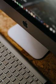 A close-up view of an Apple iMac computer monitor and keyboard setup on a wooden surface. The focus is on the sleek design and clean lines of the Apple hardware, with the Apple logo visible on the monitor. The keyboard is an Apple Magic Keyboard with a white finish, reflecting minimal and tidy aesthetics.