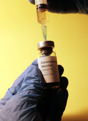 A vial labeled 'Coronavirus Vaccine COVID-19 Injection Only' is held by a gloved hand. A syringe, held by another gloved hand, is inserted into the vial. The background is a solid yellow color.