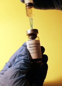 A vial labeled 'Coronavirus Vaccine COVID-19 Injection Only' is held by a gloved hand. A syringe, held by another gloved hand, is inserted into the vial. The background is a solid yellow color.