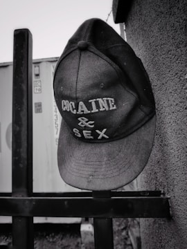 A baseball cap hangs on a metal railing, with bold lettering reading 'COCAINE & SEX'. The background consists of a textured wall and the side of a shipping container.