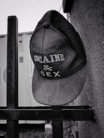 A baseball cap hangs on a metal railing, with bold lettering reading 'COCAINE & SEX'. The background consists of a textured wall and the side of a shipping container.
