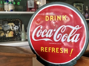 A vibrant red circular Coca-Cola sign with white and yellow text reading 'Drink Coca-Cola Refresh!' is positioned prominently in the foreground. In the background, there is a shelf displaying various vintage items such as antique bottles, a framed picture, a metal tray with decorative items, and another soda bottle.