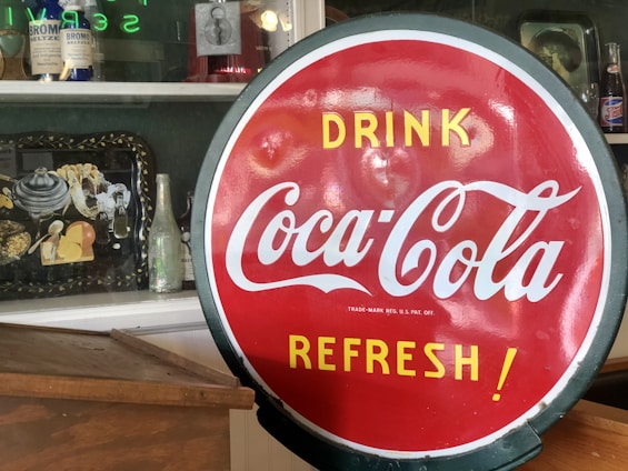 A vibrant red circular Coca-Cola sign with white and yellow text reading 'Drink Coca-Cola Refresh!' is positioned prominently in the foreground. In the background, there is a shelf displaying various vintage items such as antique bottles, a framed picture, a metal tray with decorative items, and another soda bottle.