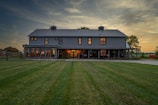 A large, two-story barn-style house with a rustic appearance stands prominently in the landscape. The structure features a gray metal roof, numerous windows, and a wrap-around porch. The warm glow of interior lights contrasts with the cool evening light. The surrounding area includes a well-maintained grassy yard, trees, and a hint of a rural setting with a visible fence and red farm equipment to the right.