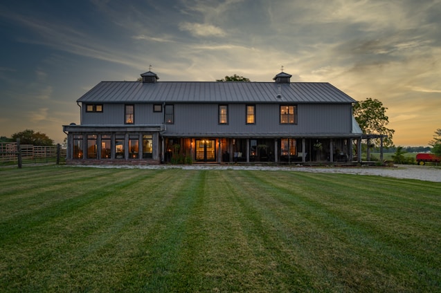 A large, two-story barn-style house with a rustic appearance stands prominently in the landscape. The structure features a gray metal roof, numerous windows, and a wrap-around porch. The warm glow of interior lights contrasts with the cool evening light. The surrounding area includes a well-maintained grassy yard, trees, and a hint of a rural setting with a visible fence and red farm equipment to the right.