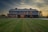A large, two-story barn-style house with a rustic appearance stands prominently in the landscape. The structure features a gray metal roof, numerous windows, and a wrap-around porch. The warm glow of interior lights contrasts with the cool evening light. The surrounding area includes a well-maintained grassy yard, trees, and a hint of a rural setting with a visible fence and red farm equipment to the right.