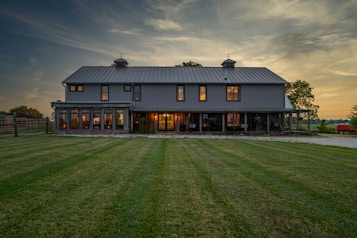 A large, two-story barn-style house with a rustic appearance stands prominently in the landscape. The structure features a gray metal roof, numerous windows, and a wrap-around porch. The warm glow of interior lights contrasts with the cool evening light. The surrounding area includes a well-maintained grassy yard, trees, and a hint of a rural setting with a visible fence and red farm equipment to the right.