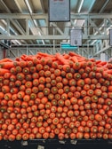 A large, tidy stack of bright orange carrots is displayed prominently in a market setting. Each carrot is uniformly sized and neatly piled, showcasing their orange tips and green tops. Overhead, there is signage displaying pricing details, and the background features a high warehouse-like ceiling with exposed beams.