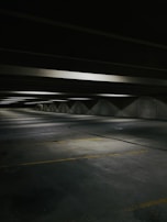 A dimly lit, empty parking garage with concrete floors and ceilings. The space is lined with fluorescent lights casting soft shadows. Yellow parking lines mark the parking spaces on the floor.