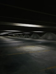 A dimly lit, empty parking garage with concrete floors and ceilings. The space is lined with fluorescent lights casting soft shadows. Yellow parking lines mark the parking spaces on the floor.