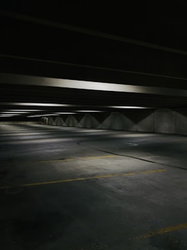 A dimly lit, empty parking garage with concrete floors and ceilings. The space is lined with fluorescent lights casting soft shadows. Yellow parking lines mark the parking spaces on the floor.