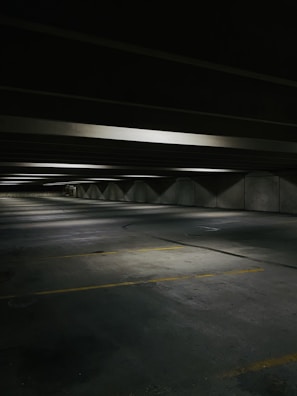 A dimly lit, empty parking garage with concrete floors and ceilings. The space is lined with fluorescent lights casting soft shadows. Yellow parking lines mark the parking spaces on the floor.