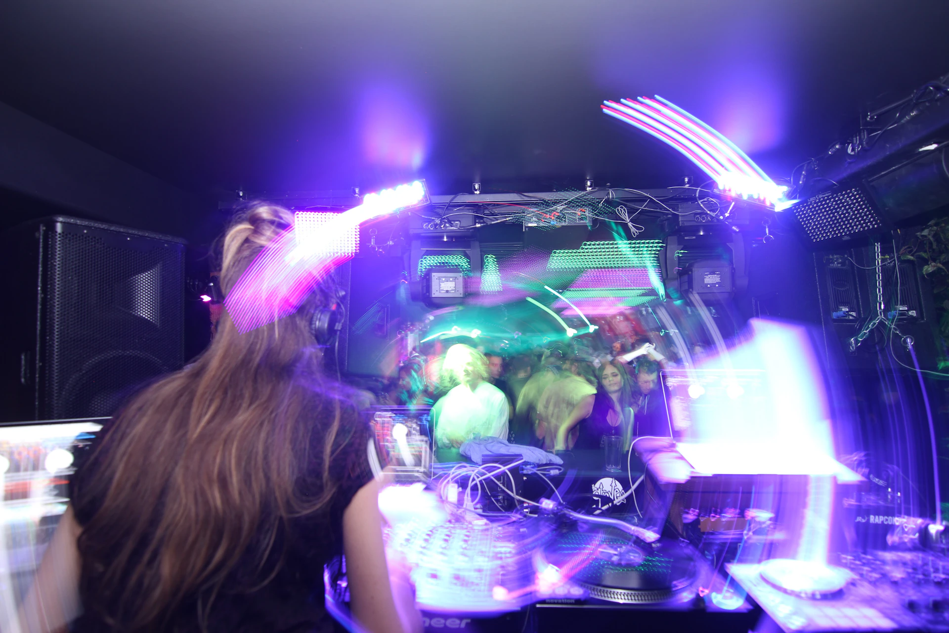 A person with long hair is in a dark room with vibrant neon lighting, standing in front of DJ equipment, including a turntable. The room is filled with colorful light trails and reflections, with people in the background, creating a lively and energetic atmosphere.