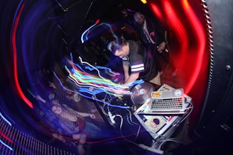 A DJ is performing in a dimly lit club, surrounded by colorful swirling lights. The setup includes turntables and a laptop covered with stickers. A crowd is enthusiastically dancing, with some people raising their hands in the air.