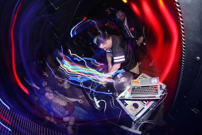 A DJ is performing in a dimly lit club, surrounded by colorful swirling lights. The setup includes turntables and a laptop covered with stickers. A crowd is enthusiastically dancing, with some people raising their hands in the air.
