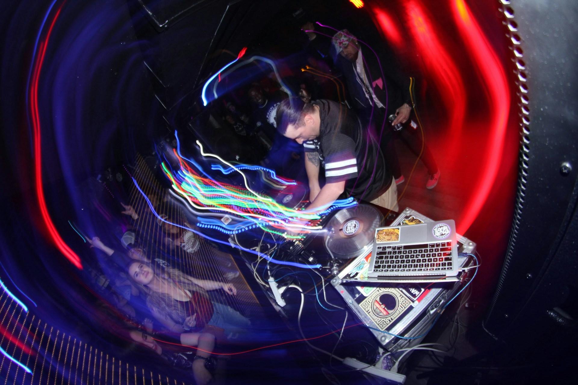 A DJ is performing in a dimly lit club, surrounded by colorful swirling lights. The setup includes turntables and a laptop covered with stickers. A crowd is enthusiastically dancing, with some people raising their hands in the air.
