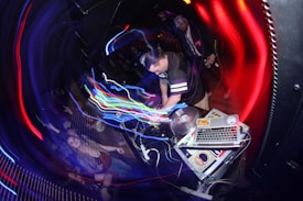 A DJ is performing in a dimly lit club, surrounded by colorful swirling lights. The setup includes turntables and a laptop covered with stickers. A crowd is enthusiastically dancing, with some people raising their hands in the air.