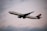 A commercial airplane is flying through the sky, showcasing its large wings and engines. The aircraft is white with a blue tail fin marked with a logo. The backdrop is a cloudy sky with soft lighting, suggesting it might be either early morning or late evening.