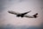 A commercial airplane is flying through the sky, showcasing its large wings and engines. The aircraft is white with a blue tail fin marked with a logo. The backdrop is a cloudy sky with soft lighting, suggesting it might be either early morning or late evening.