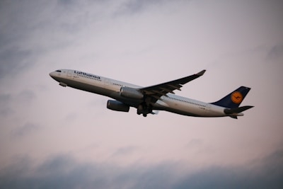 A commercial airplane is flying through the sky, showcasing its large wings and engines. The aircraft is white with a blue tail fin marked with a logo. The backdrop is a cloudy sky with soft lighting, suggesting it might be either early morning or late evening.