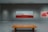 A modern art gallery features a large painting with a vivid red foreground and a muted mountainous background. The room's walls are a neutral shade, and a wooden bench sits in front of the artwork on a polished concrete floor.