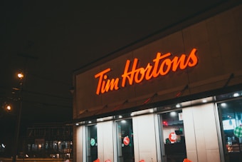 A well-lit Tim Hortons café displays its bright neon signage against a dimly lit background. The interior lights of the café are visible through the large windows, which are decorated with festive stickers. The scene captures the street outside, with some streetlights adding a warm glow.