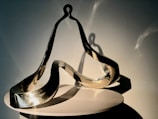 A modern, abstract metal sculpture with smooth, flowing curves and reflective surfaces sits on a circular white pedestal. The sculpture is lit from one side, casting intricate shadows and highlights that emphasize its dynamic form.