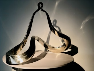 A modern, abstract metal sculpture with smooth, flowing curves and reflective surfaces sits on a circular white pedestal. The sculpture is lit from one side, casting intricate shadows and highlights that emphasize its dynamic form.