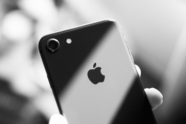 An Apple iPhone is held in someone's hand. The focus is on the back of the phone, featuring the Apple logo. The image is in black and white, with a focus on the sleek and reflective surface of the device.