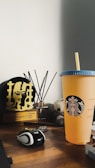 A wooden desk displaying an assortment of objects including a yellow-orange Starbucks cup with a blue lid, a black and white computer mouse, a trophy with a plaque, a fragrance diffuser with black sticks, and a small stuffed toy panda. The background is a plain white wall.