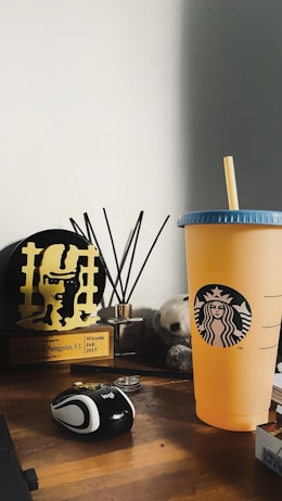 A wooden desk displaying an assortment of objects including a yellow-orange Starbucks cup with a blue lid, a black and white computer mouse, a trophy with a plaque, a fragrance diffuser with black sticks, and a small stuffed toy panda. The background is a plain white wall.