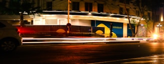 Nighttime urban scene with bright street lights creating light trails from passing cars. A partially illuminated building has signs indicating something is 'coming soon.' Trees are visible next to the road, and the atmosphere suggests a bustling city environment with a mix of artificial and natural elements.